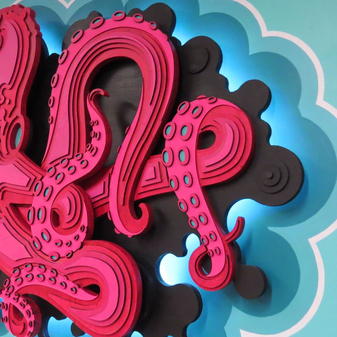 A vibrant fuchsia 3D layered wood octopus floats against a black ink backdrop, set off by a turquoise background and illuminated by LED lighting for a striking visual effect.