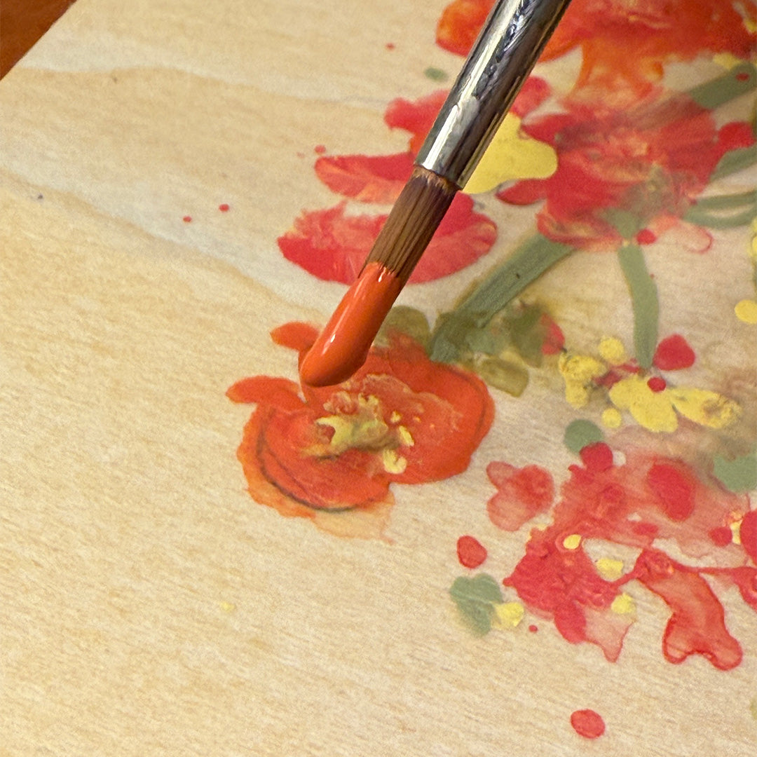 Paintbrush with orange paint on a floral painting on authentic wood grain panel.