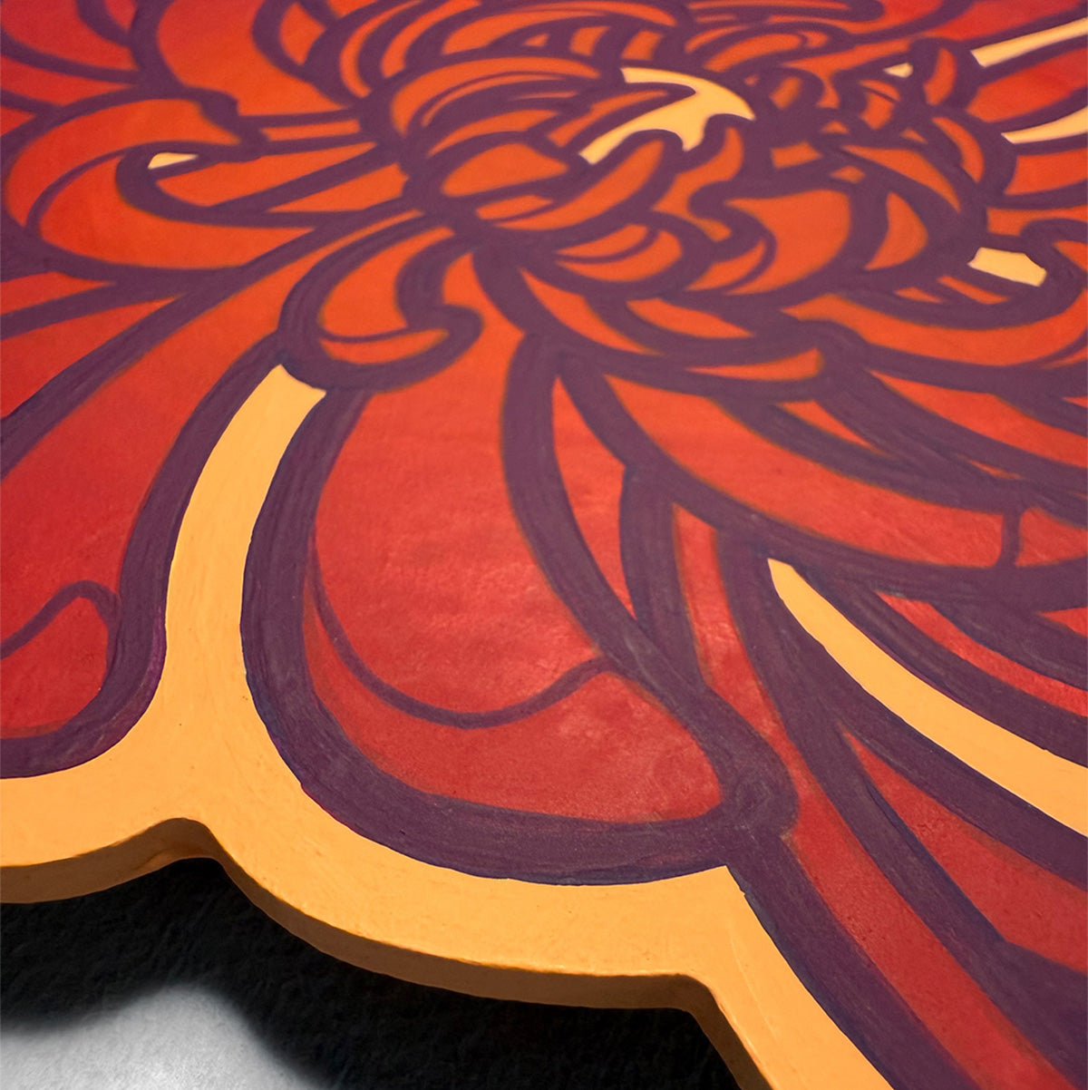 A close-up of a decorative wooden painting featuring a stylized floral design in red, orange, and dark purple, with a scalloped edge and visible wood thickness.