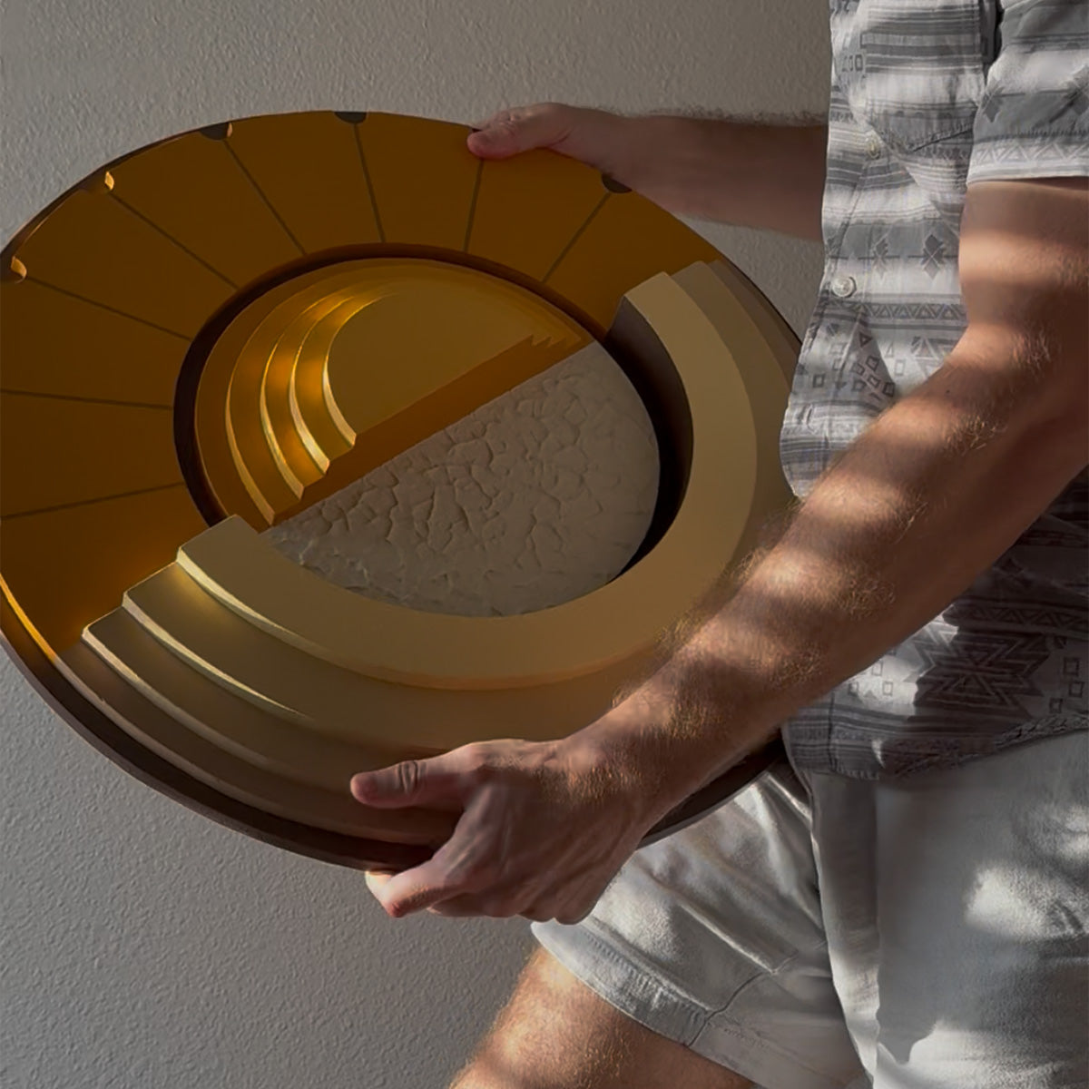 A person holding a large, circular, tiered wood wall art in warm mustard yellow and brown tones against a neutral wall.