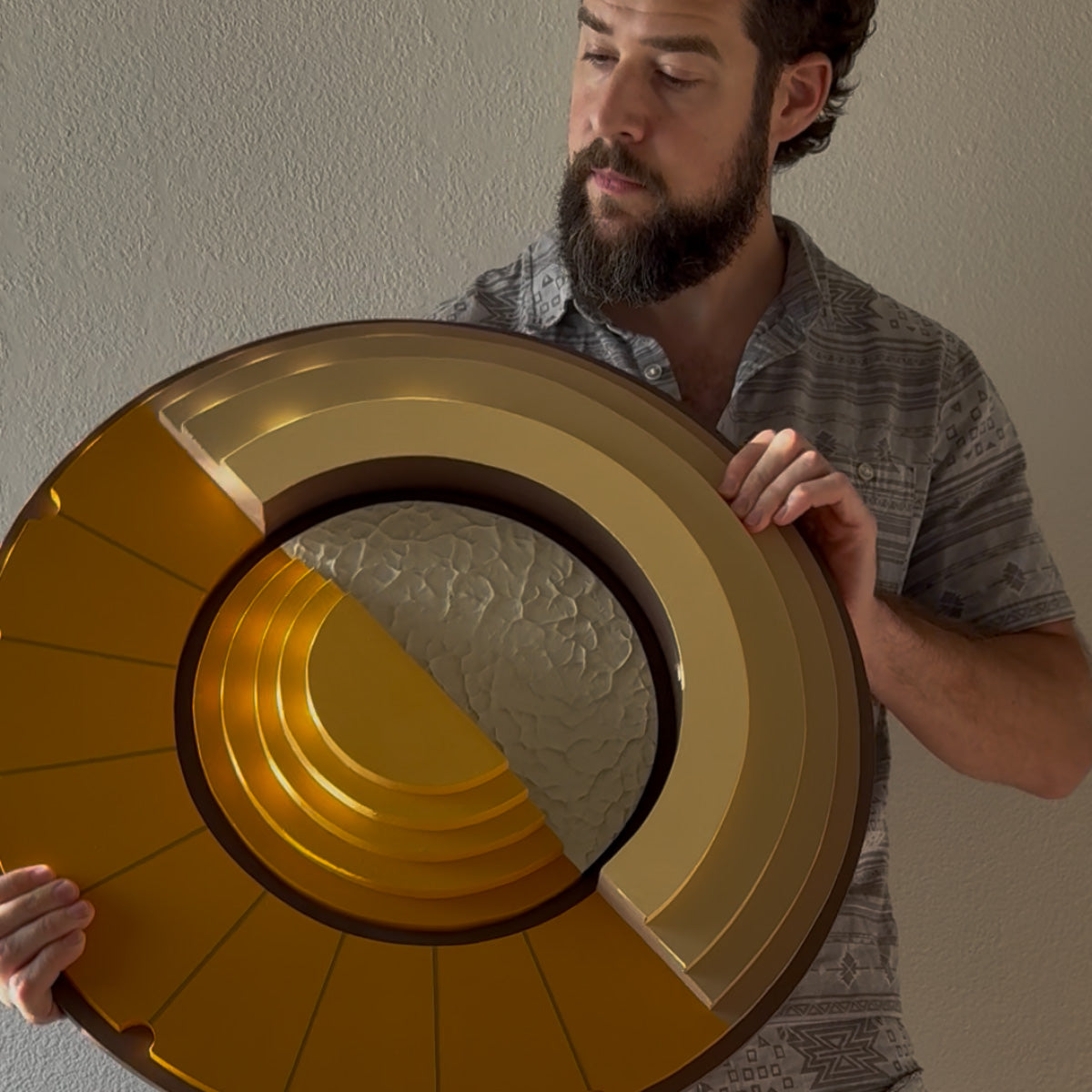 A man with a beard holds a large circular layered wood wall art with a yellow and brown finish and a textured cream center, against a plain beige wall.