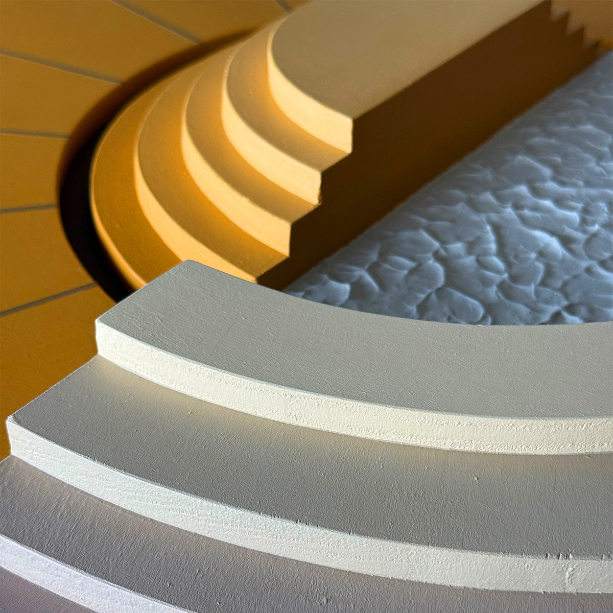 Close-up of a modern architectural wood wall art with curved beige layered steps in the foreground, a textured light gray surface, and warm yellow geometric forms in the background.