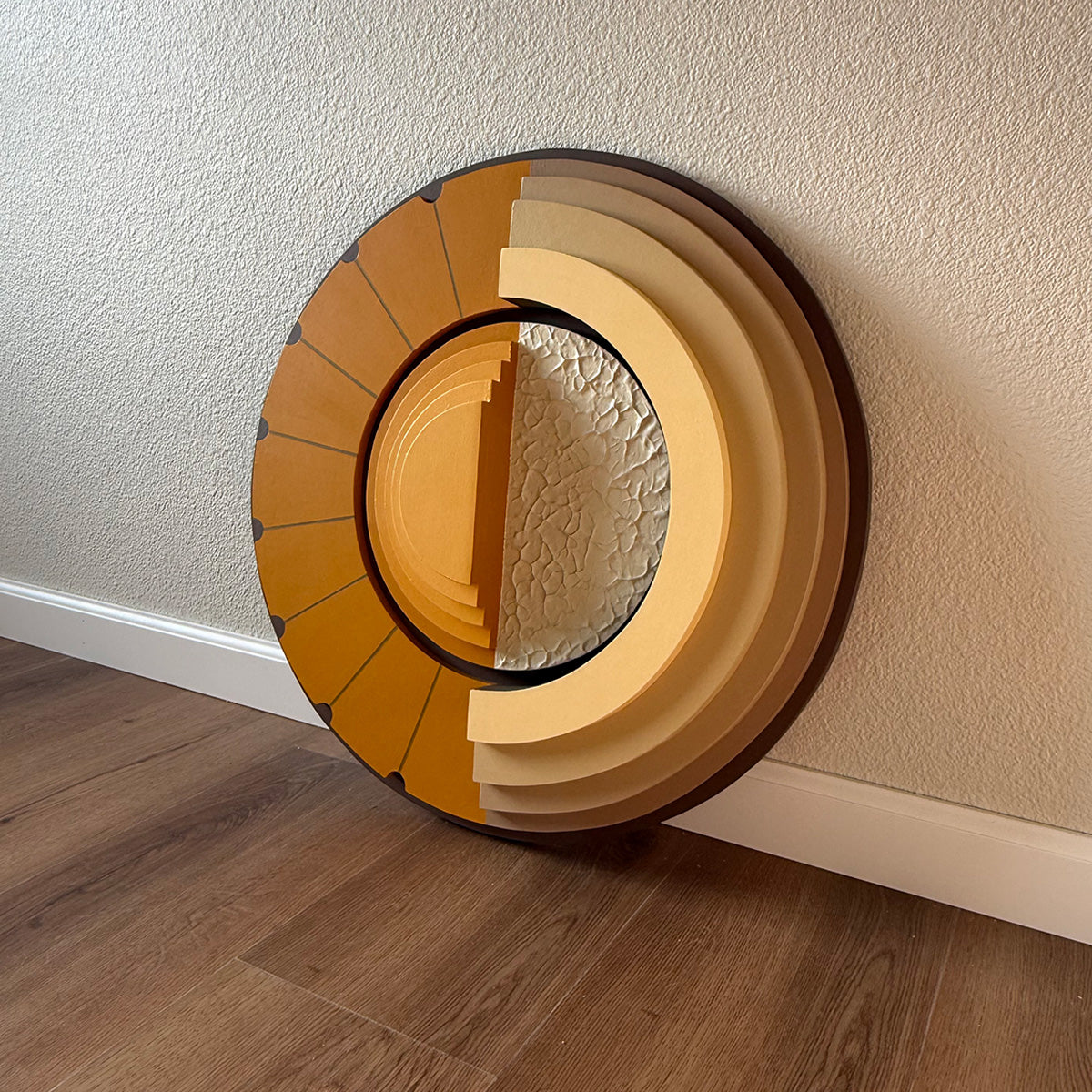 A large circular wood wall art with concentric rings, leaning against a textured beige wall on a warm brown wood floor.