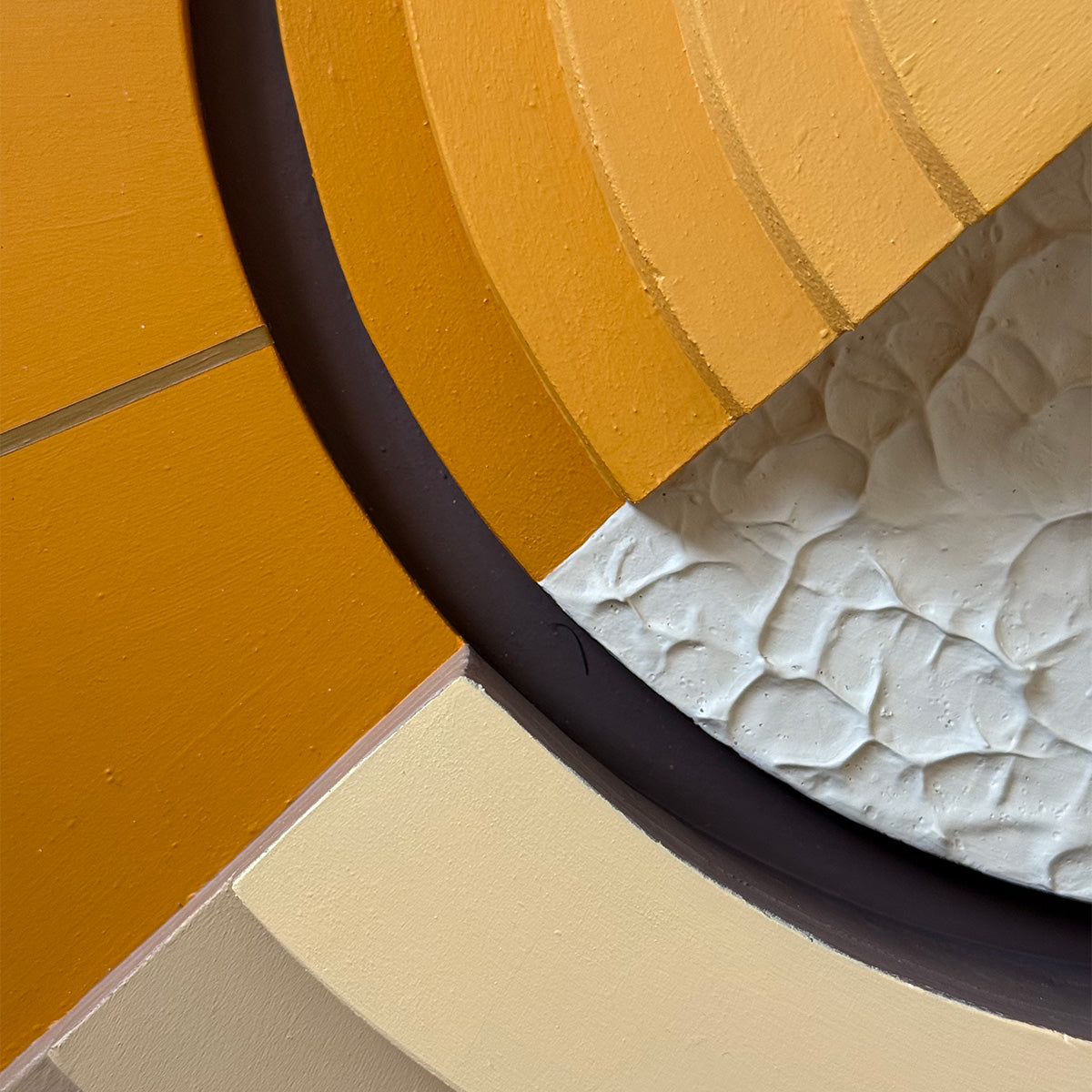 Abstract close-up of a textured circular wall art design with mustard yellow, cream, and textured organic shapes separated by a dark curved accent line.
