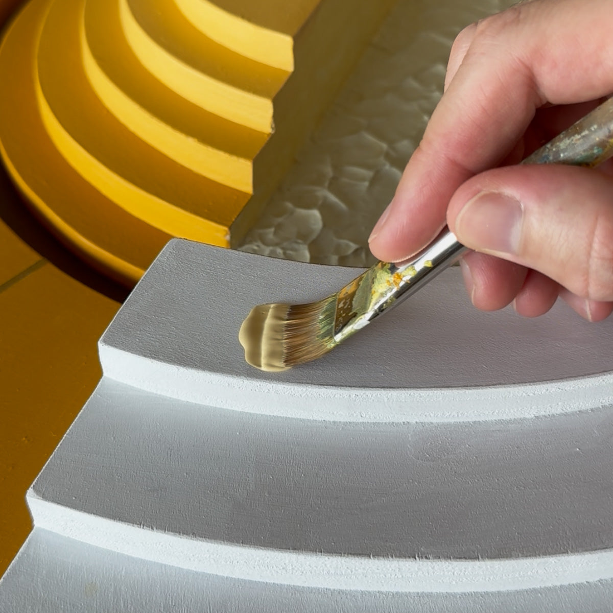 A close-up of a hand holding a paintbrush and applying light yellow paint to a primed white curved step, with a yellow layered background and textured surface visible.