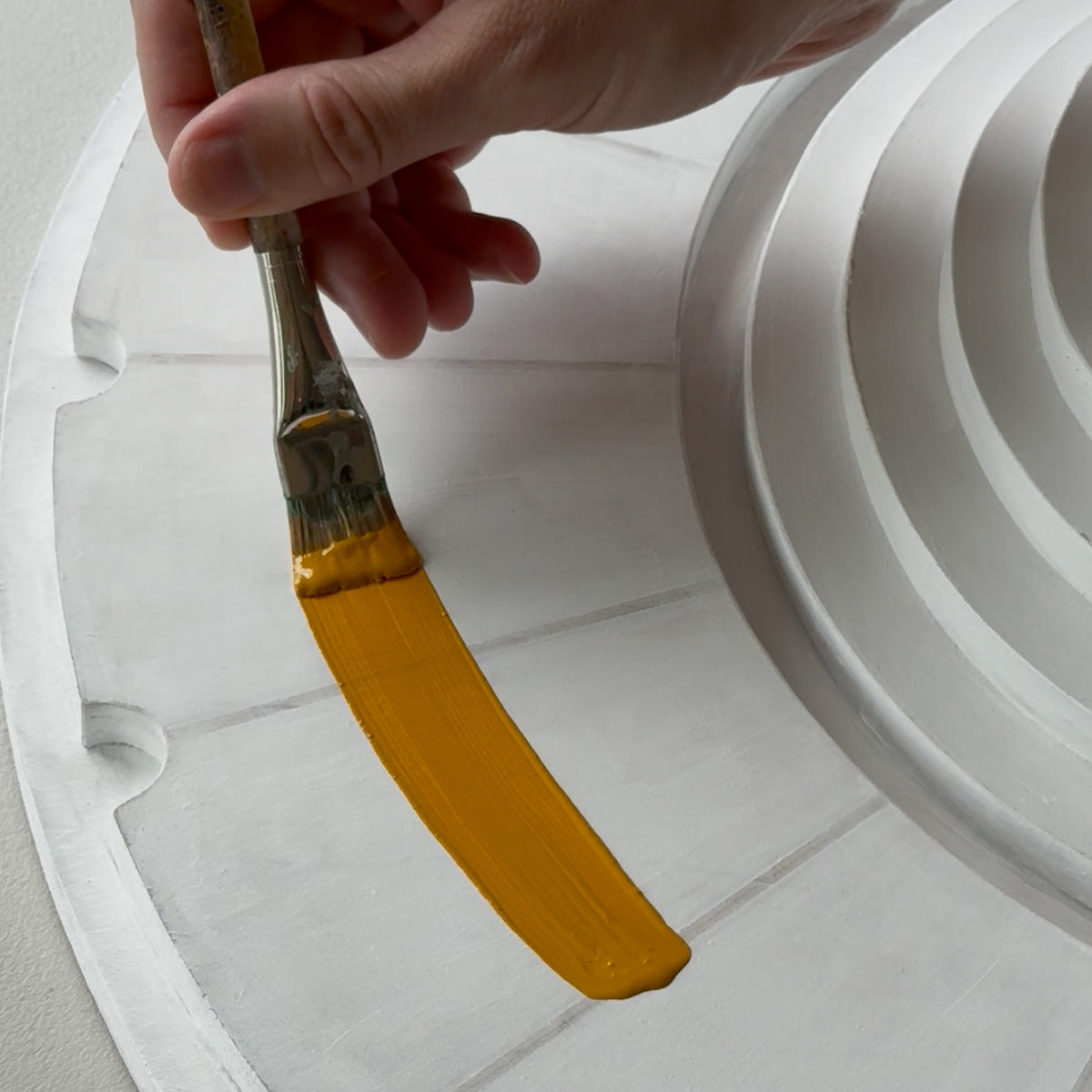 A hand holding a paintbrush applies a thick coat of mustard yellow paint to a white primed circular wood piece with concentric circular ridges.