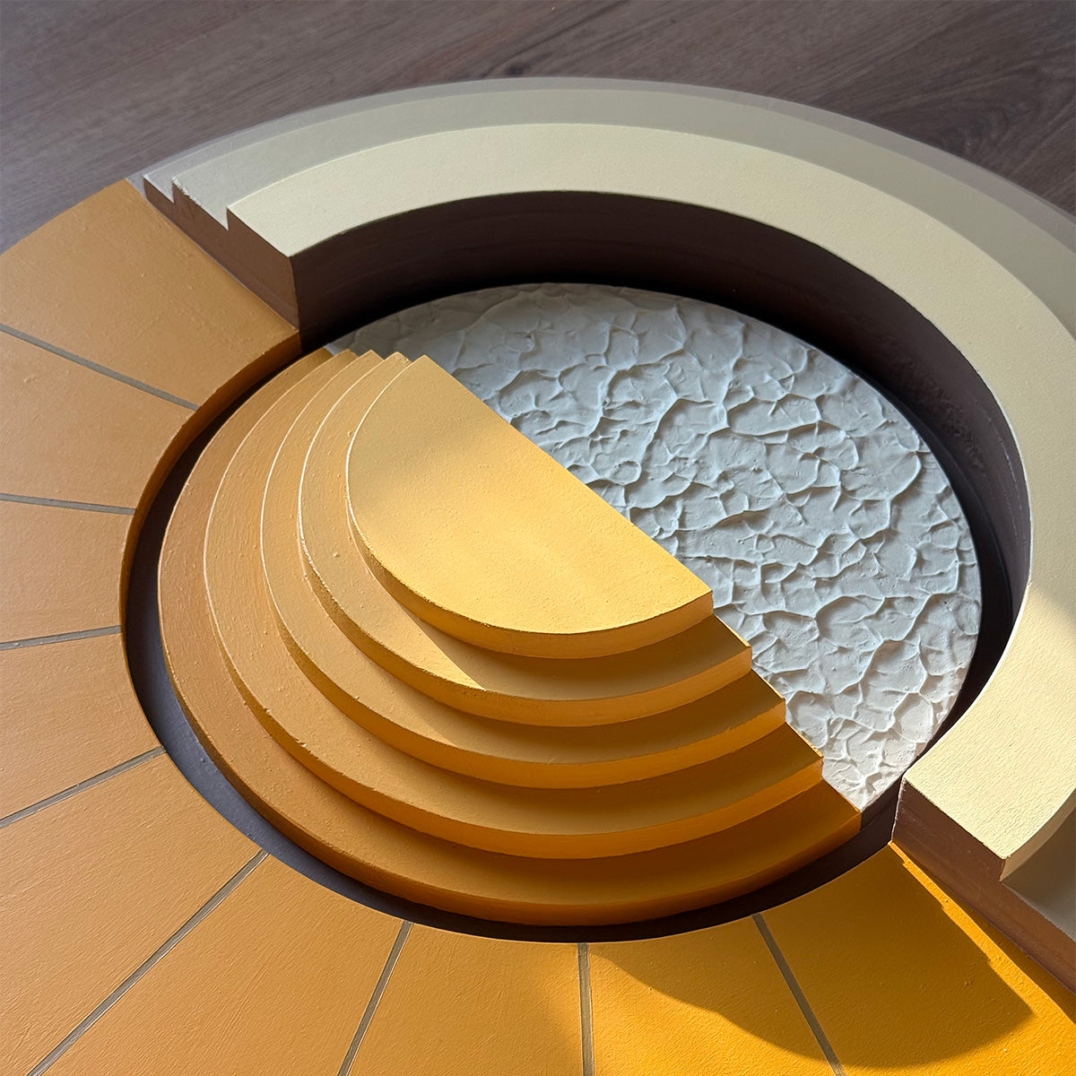A close-up of a circular layered wood art piece with concentric mustard yellow and beige rings, a textured light gray half moon, and a dark brown rim, photographed on a dark wood surface.