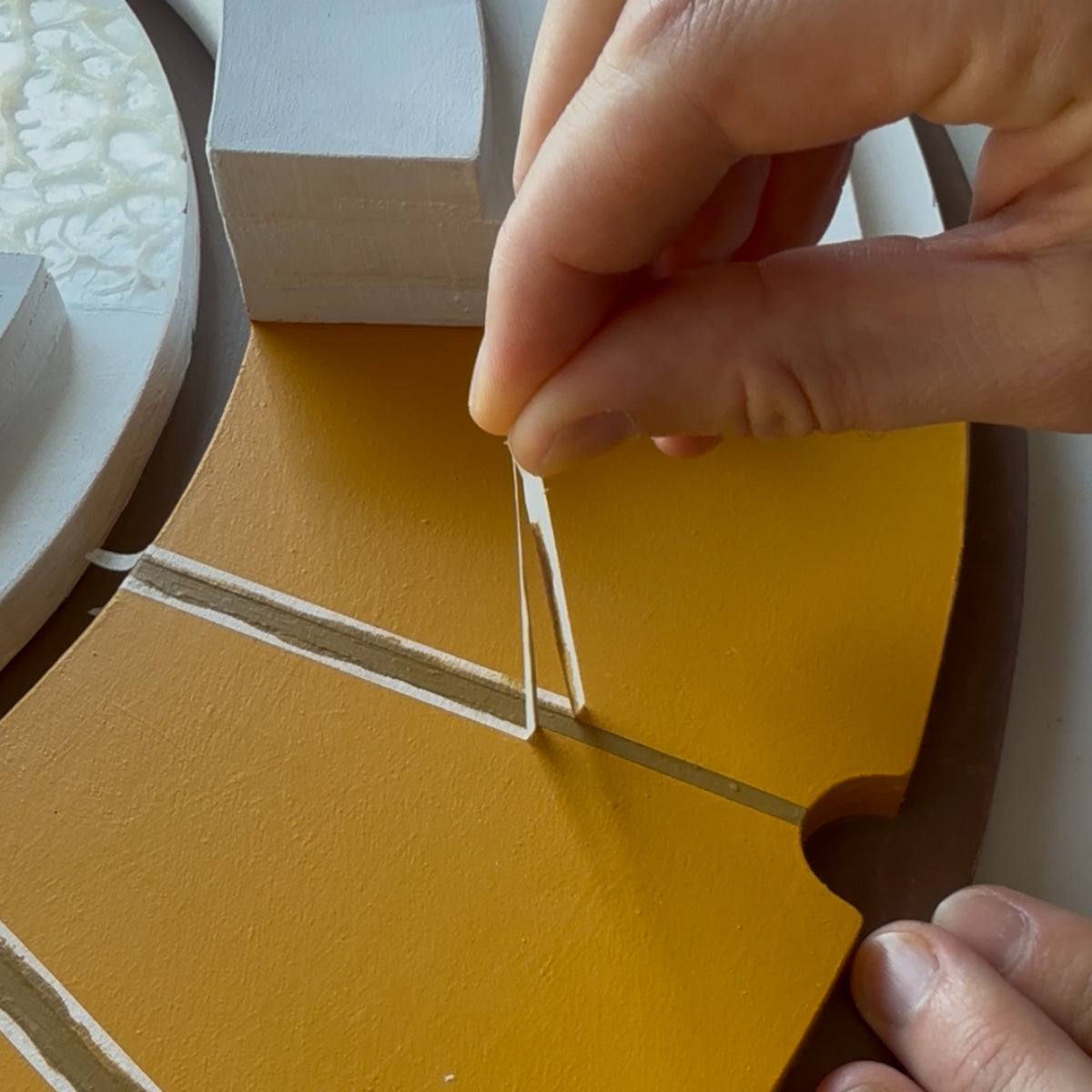 A close-up of a hand applying a thin painter's tape strip to a curved, mustard-yellow wooden panel with a brown-toned accent line.