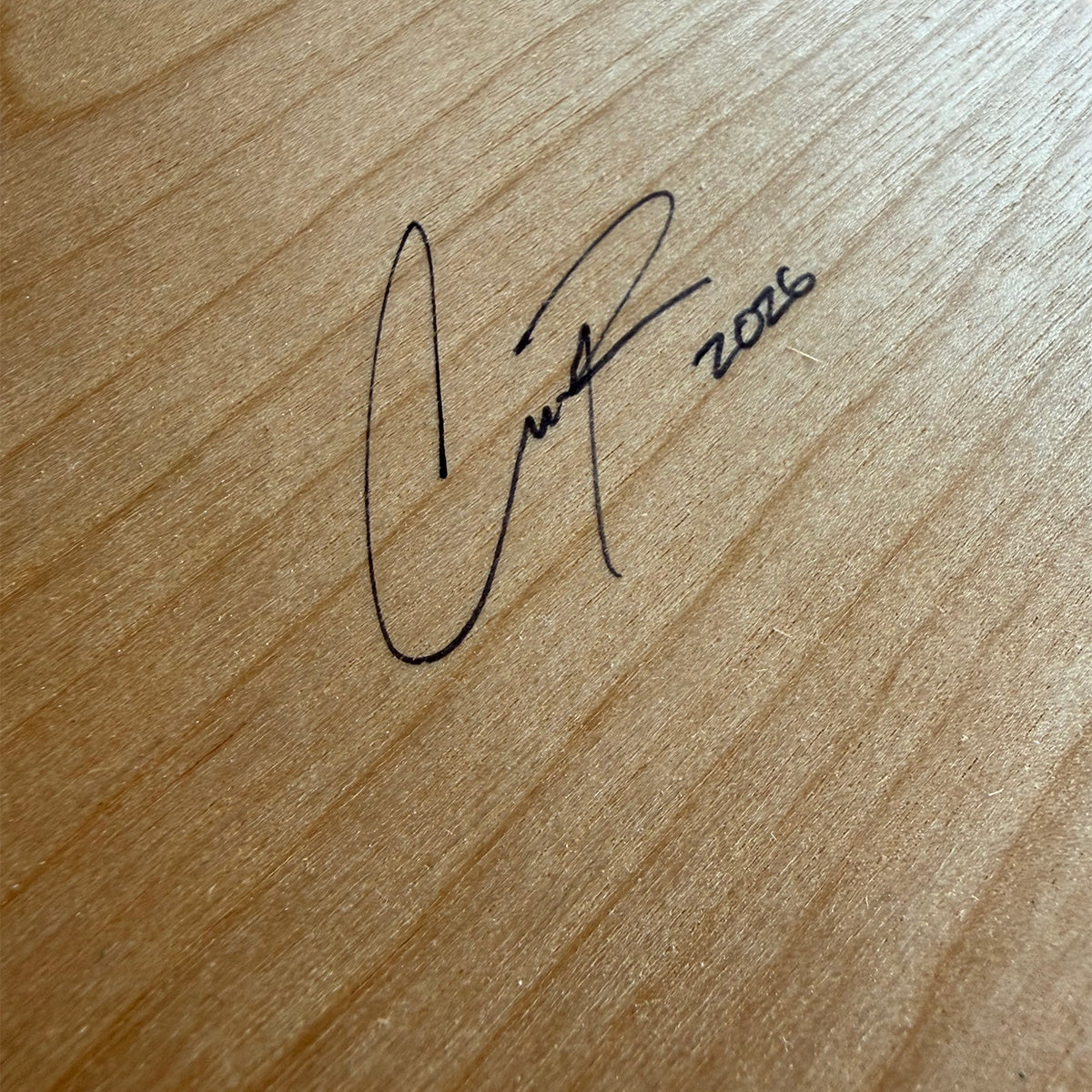 A close-up of a light wood surface with a black cursive signature and the year 2026 written near the center.