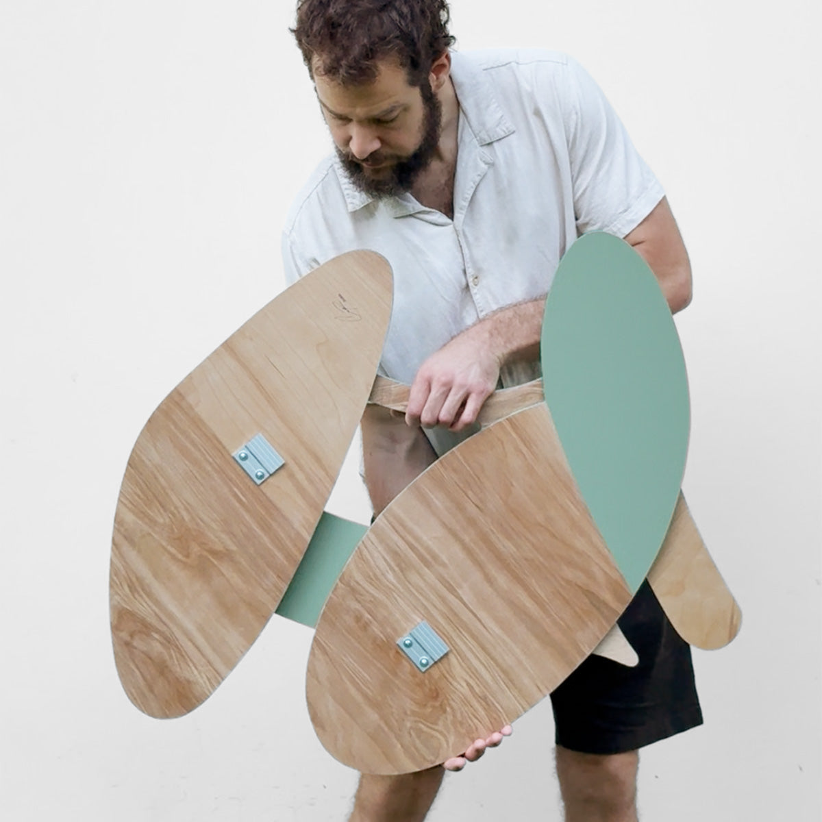 A man in a light-colored shirt and black shorts holds a modern wood art piece with large, organic-shaped wooden panels in a natural wood finish and mint green accents, set against a plain light gray background.