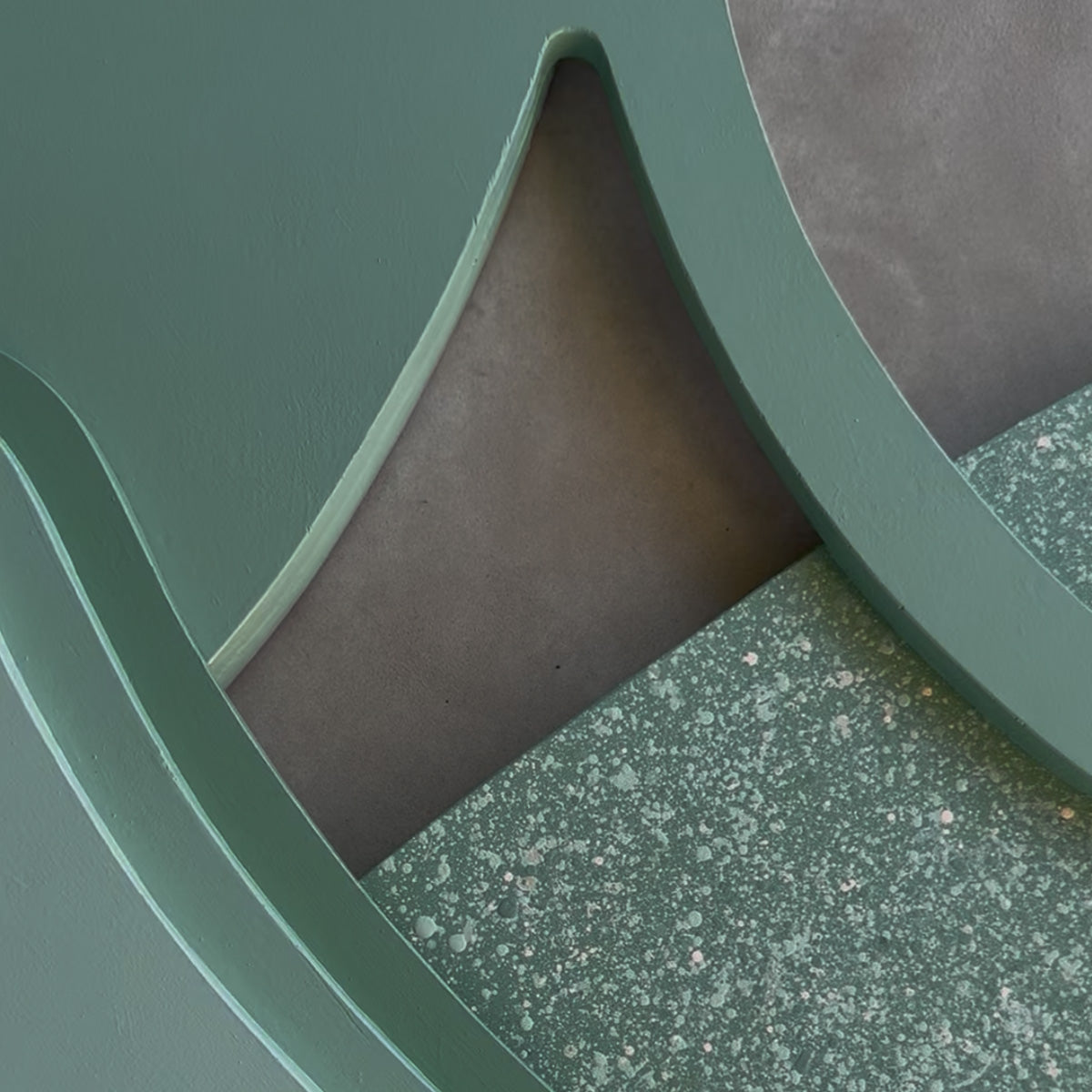 Close-up detail of a modern layered wood art showing a sage green curved shape, speckled green tones, and a gray concrete backdrop.
