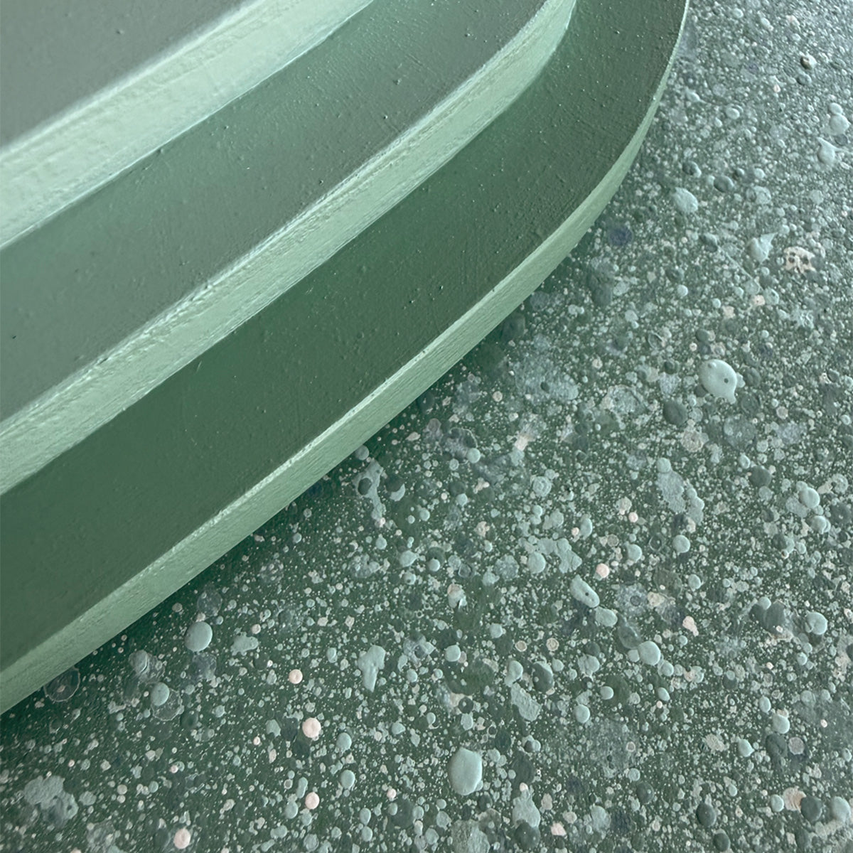 Close-up of a curved layered wood surface in a muted sage green color, shown against a speckled gray-green textured surface.