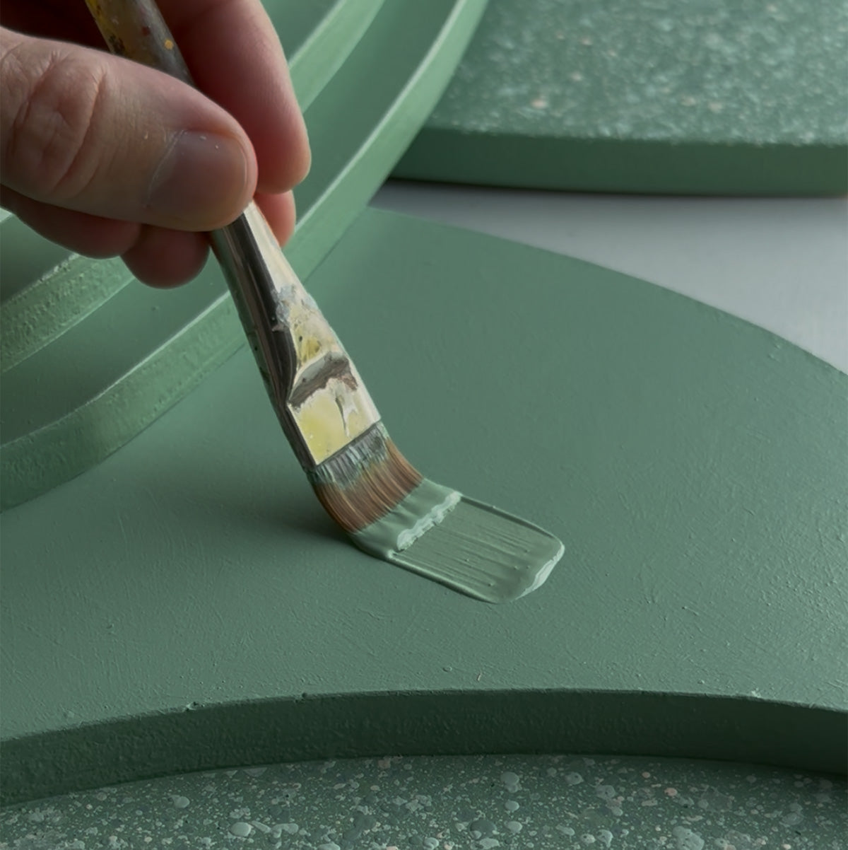 A close-up of a hand holding a paintbrush and applying a light sage green coat to a matching sage green painted surface, with textured green wood layers in the background.