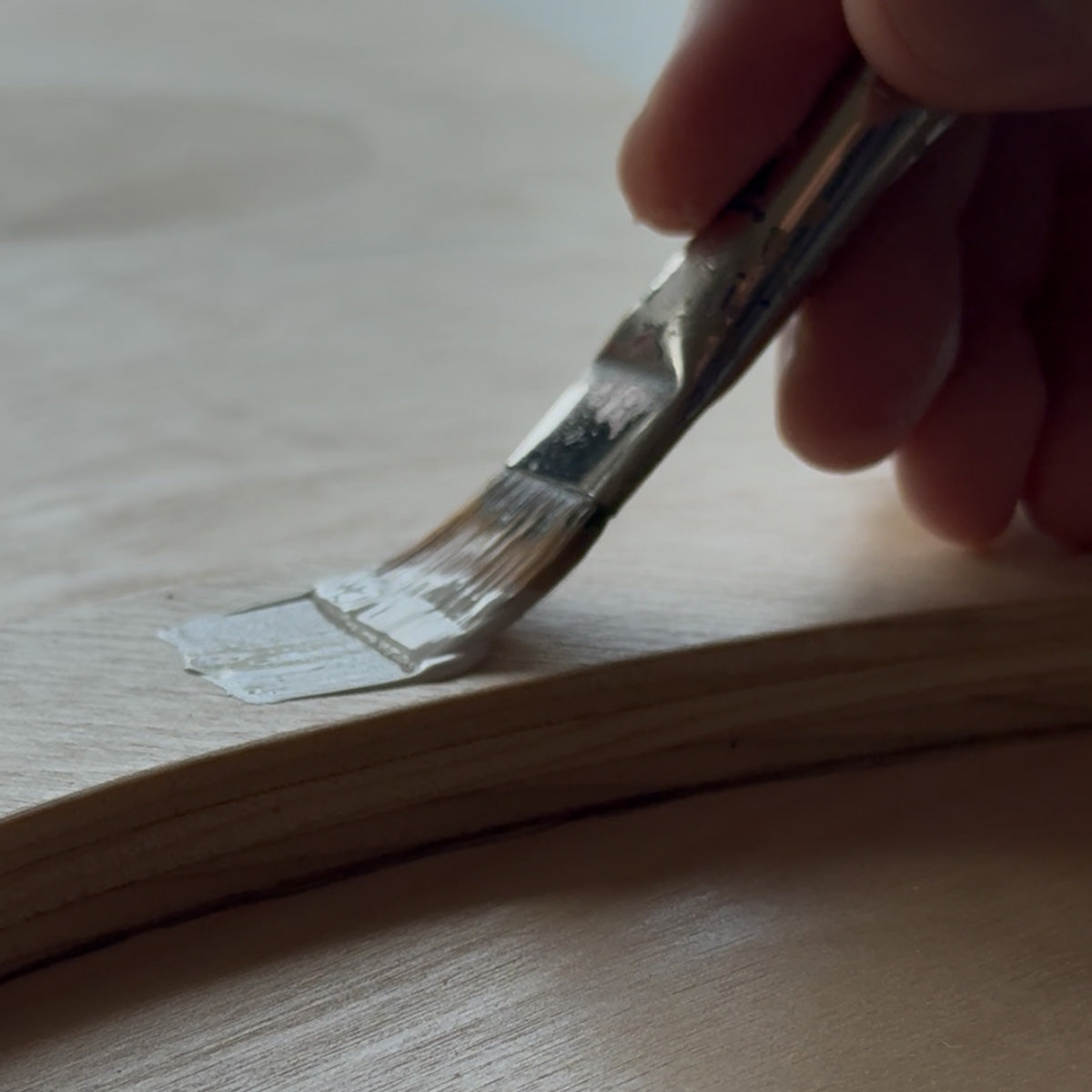 A close-up of a hand holding a paintbrush and applying a layer of white primer along the edge of a curved wooden board.