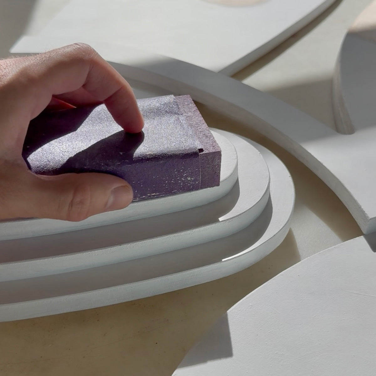 A hand holds a rectangular purple sanding block against a set of white curved geometric platforms in bright natural light.