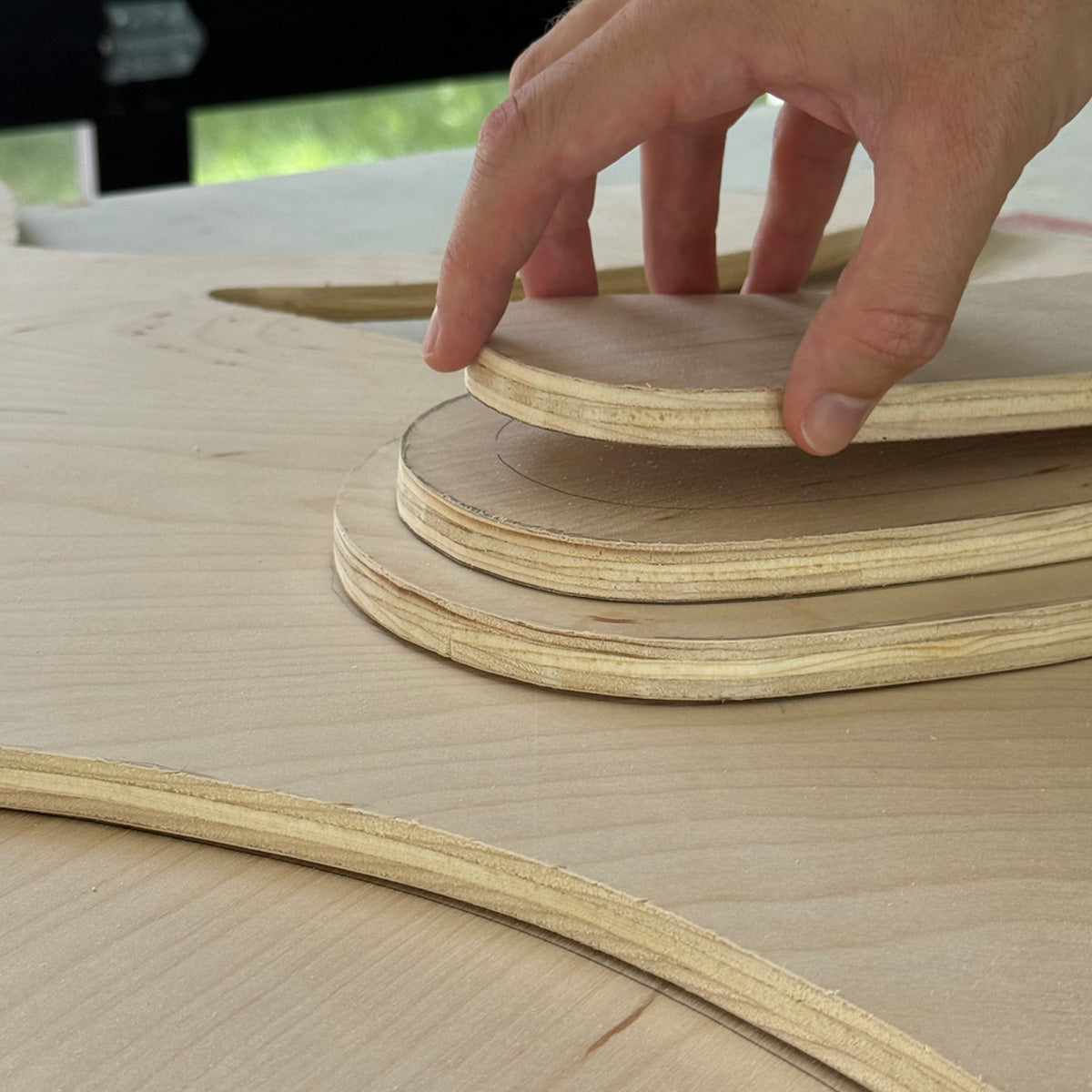 A close-up of a person's hand stacking curved, layered wood pieces on a raw wood surface.