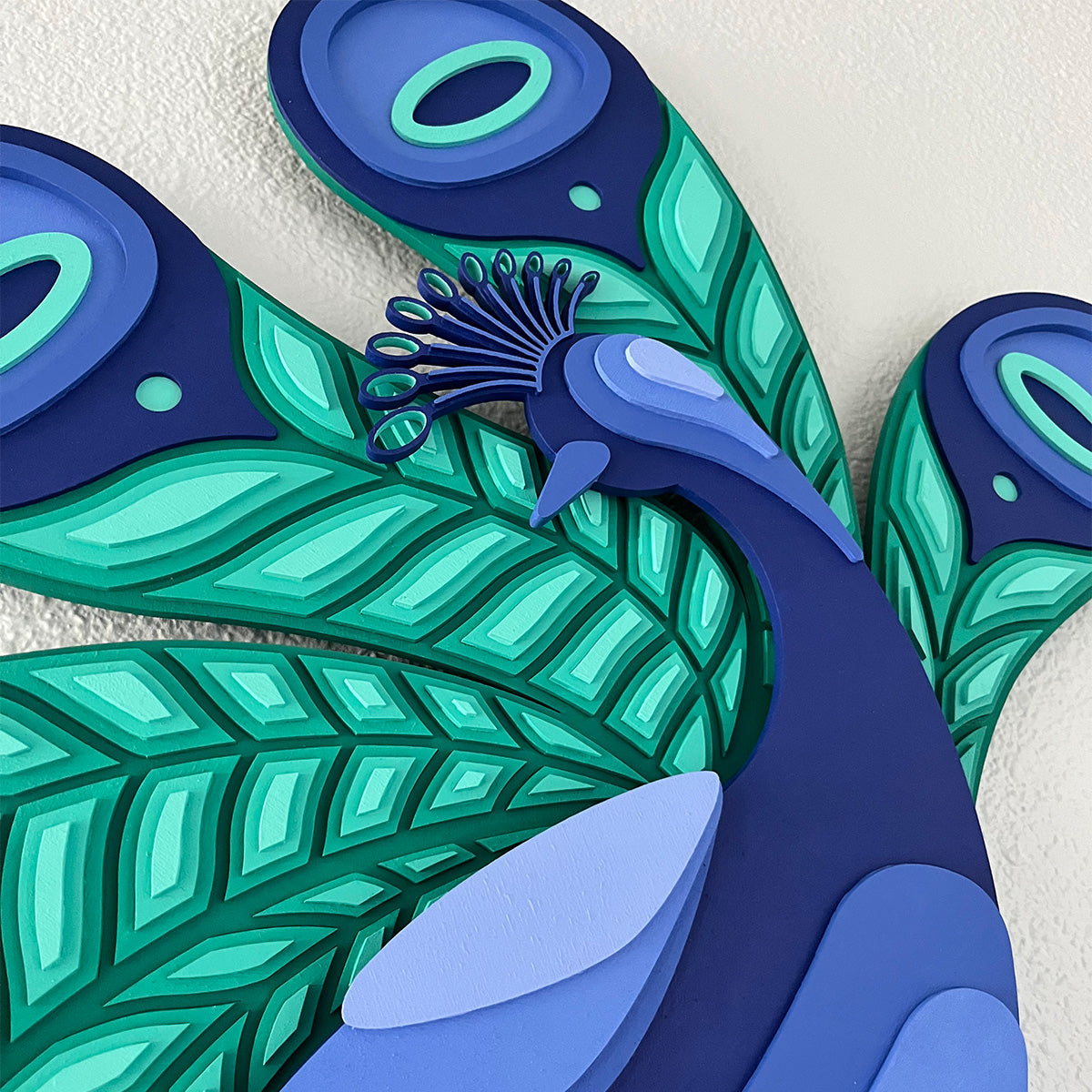 Details of a stylized peacock wall art piece made of layered wood, featuring bold blue and teal feather patterns against a white wall.