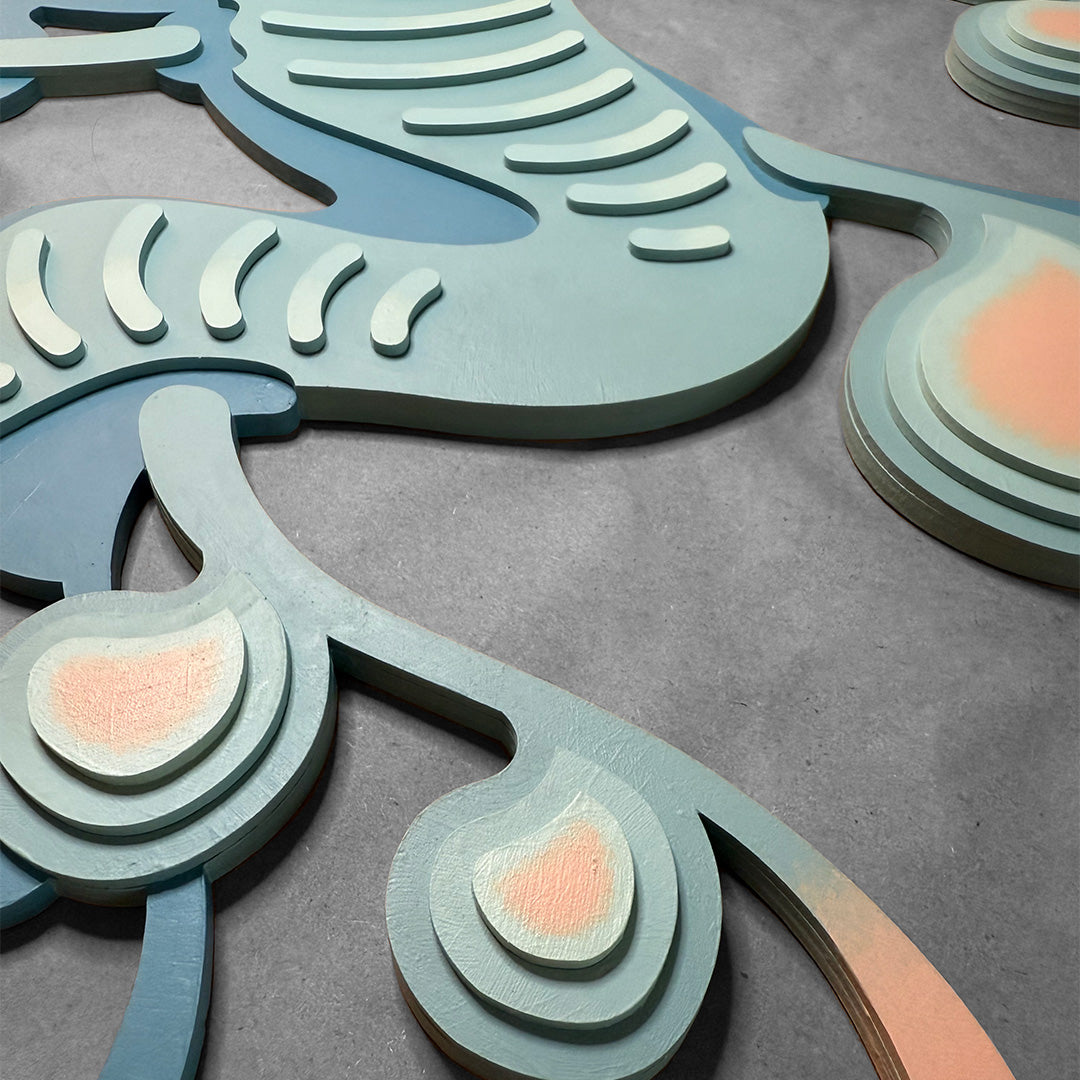 Close-up detailed photo of layered painted blue wood art with pastel pink color accents on a gray surface.