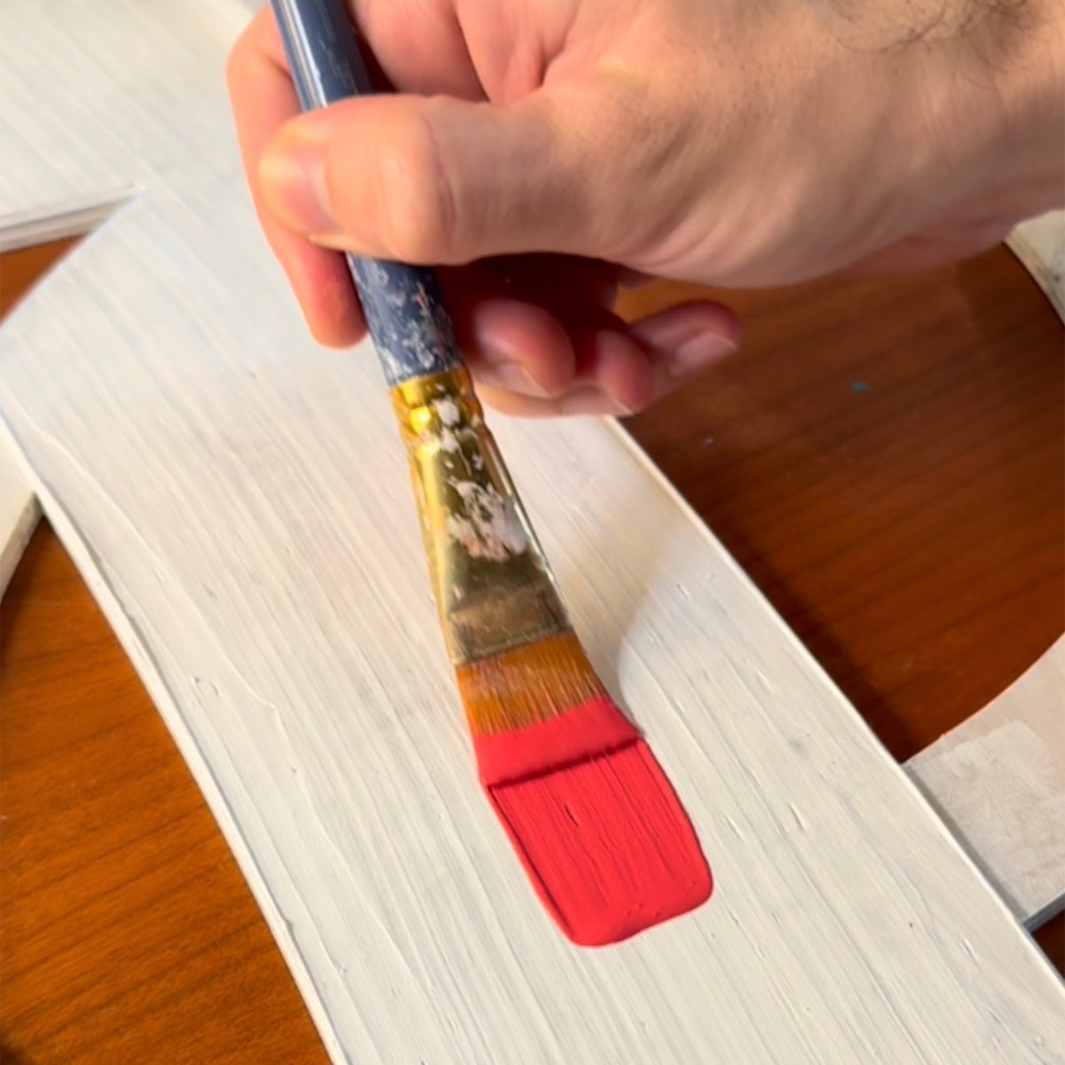 A hand holding a flat paint brush with fluorescent pink paint on the bristle head, applying the paint to a white primer acrylic texture.