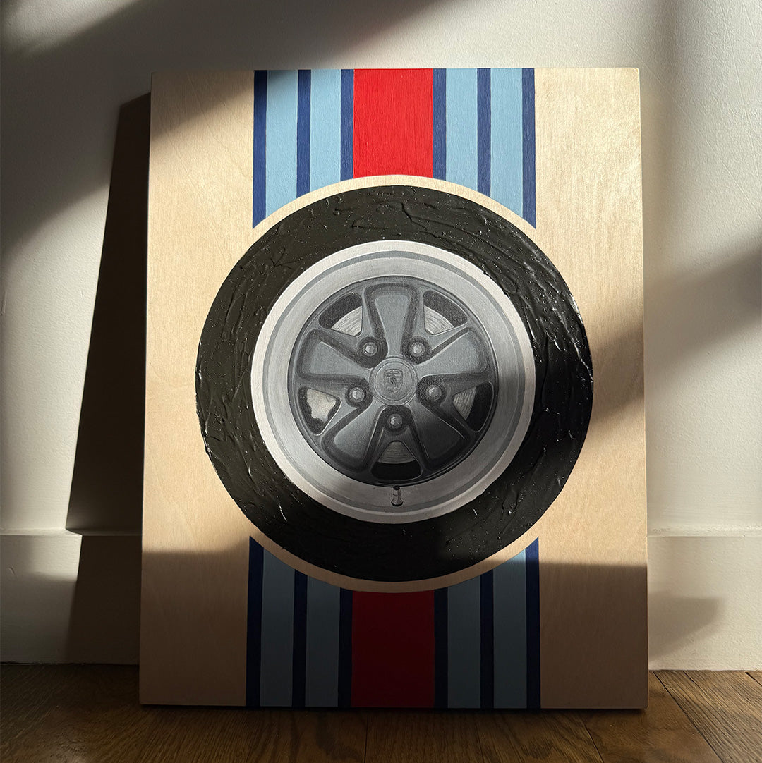 Hand-painted black porsche fuchs wheel wood wall art with textured tire and martini racing stripes behind the wheel. Laying against a wall with dramatic light and shadows across it.