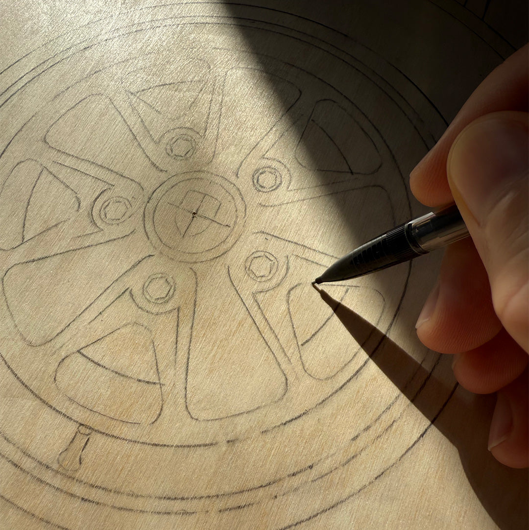 Hand holding a pencil over a wooden surface with sketch of car wheel design