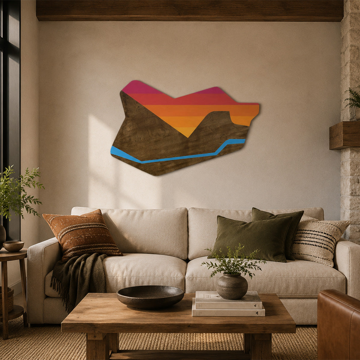Handcrafted abstract mountain sunset wood wall art displayed above a sofa in a modern rustic living room. The contour-cut design features layered sunset colors, natural wood grain mountains, and a blue river accent, illuminated by warm natural light in a cozy ranch-inspired space.